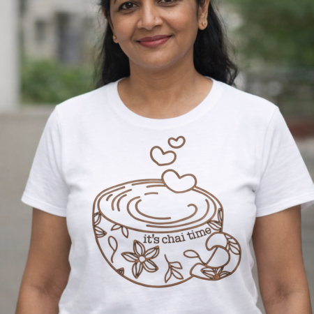 An Indian woman in her 40s stands outdoors wearing a white T-shirt with a brown line-art design of a teacup and the words “it’s chai time.” She smiles warmly, with short dark hair and gold stud earrings, against a softly blurred urban background.