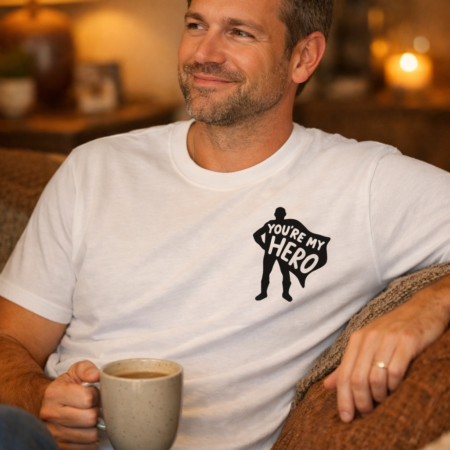 Smiling man in his late 30s wearing a white “You’re My Hero” T‑shirt, sitting on a cosy sofa with a mug in hand in a warmly lit living room.