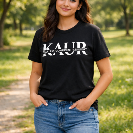 Sikh woman with light brown skin and long hair standing in a sunlit park, wearing a black T‑shirt with white KAUR text and the words ‘Fearless‑Confident‑Resilient,’ paired with blue jeans and white trainers.