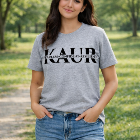 Sikh woman with light brown skin and long hair standing in a green park, wearing a heather grey KAUR T‑shirt with black text, blue jeans, and white trainers, smiling softly with hands in her pockets